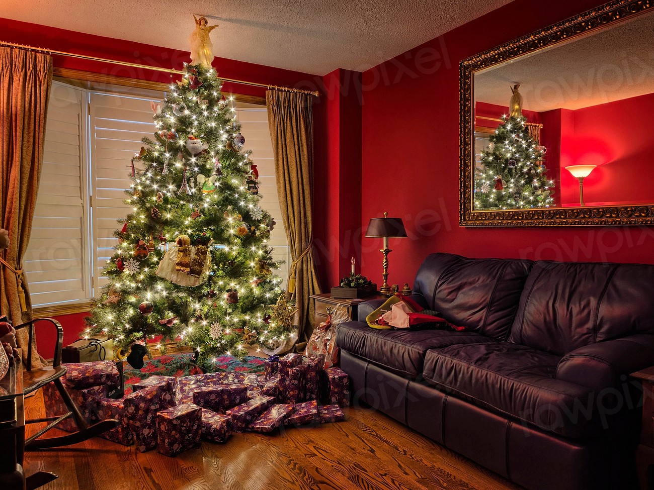 A living room where we can see a Christmas tree with presents around it. We can also see a sofa, a mirror, a window and part of a wheelchair.