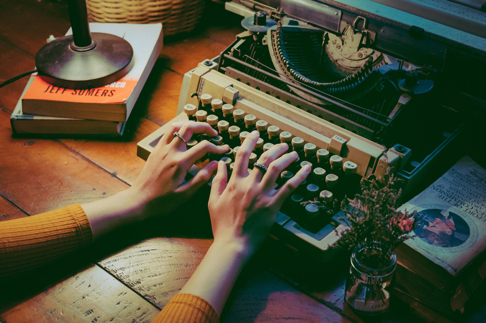 We can see a wooden desk and two hands (apparently, belonging to a woman who is notg visible in the picture) using a typewriter. There are some books on the table and flowers in a glass of water.