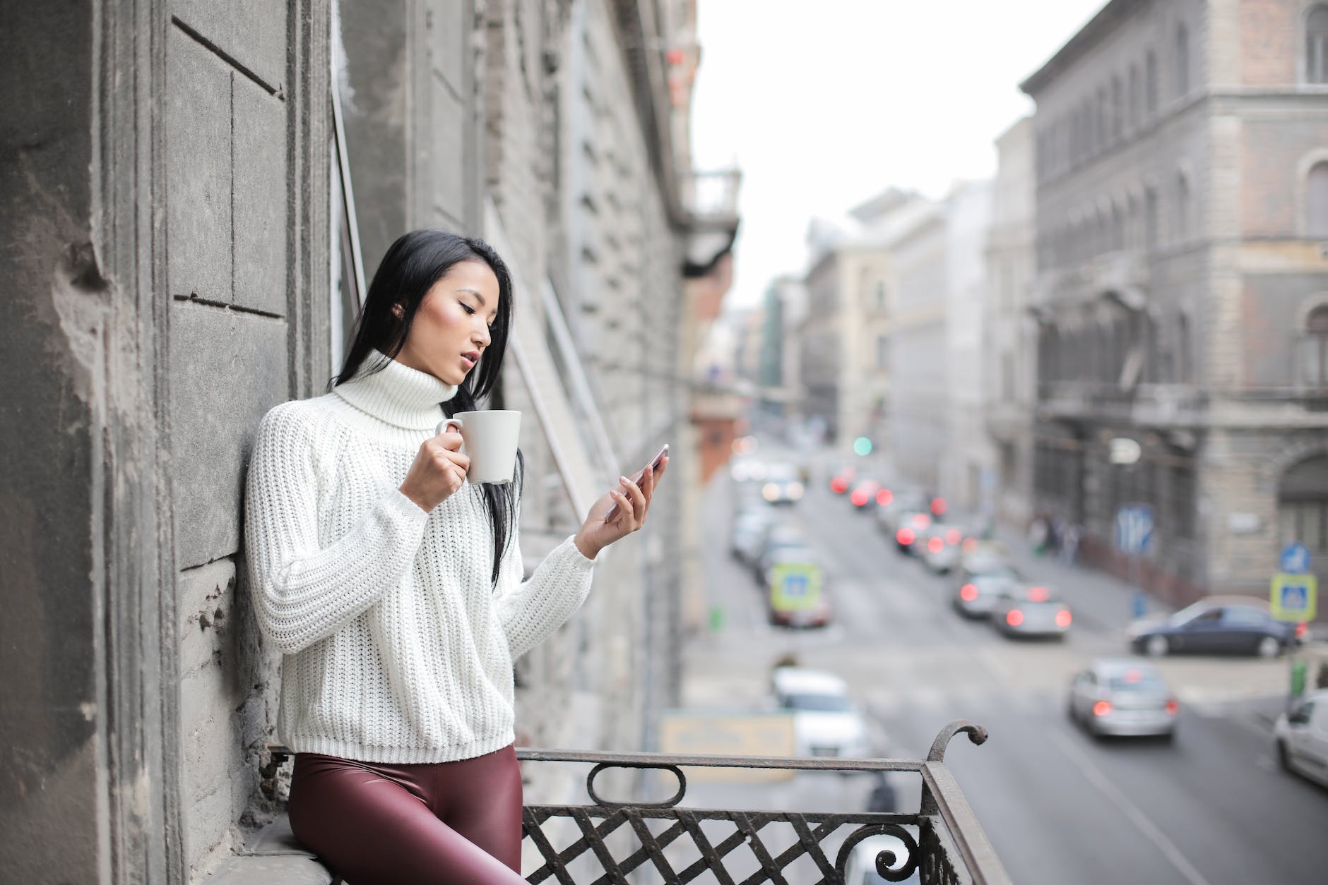 A woman is on the balcony. She has straight hair and is wearing a white pullover with turtle neck. She is also wearing purple trousers and is holding a cup of coffee. She is checking something in her cellphone.