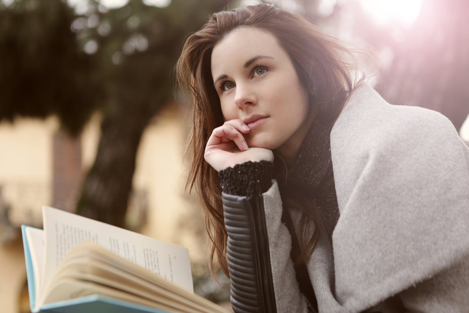 A woman is holding a book. She is outdoors. Her hair is long, straight and dark. She is wearing a gray coat and seems to be thinking about something.