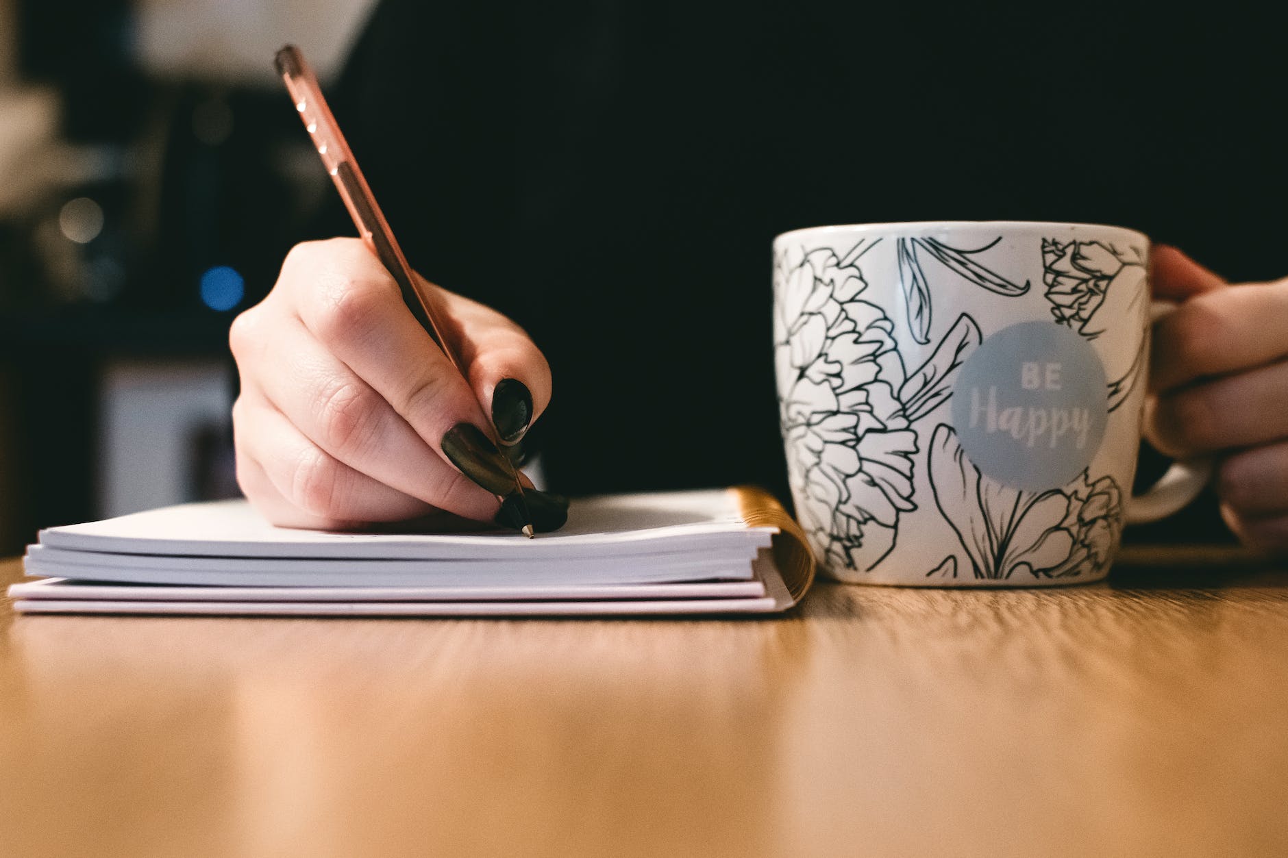A person is holding a pen while writing on a notebook. It seems to be a woman. Her nails are painted with black nail polish. She is also holding a cup of coffee where it is written "Be happy".